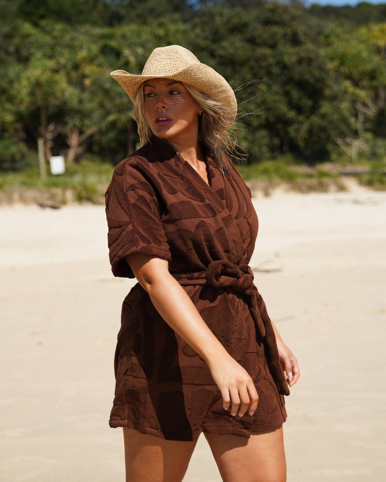 Model wearing the Ochre Lane chocolate towelling robe, front view on the beach under palm trees.