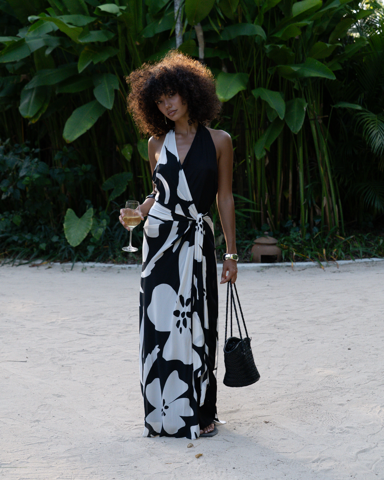 Front view of a model wearing a black and cream floral halter wrap maxi dress on a sandy garden path, holding a woven bag