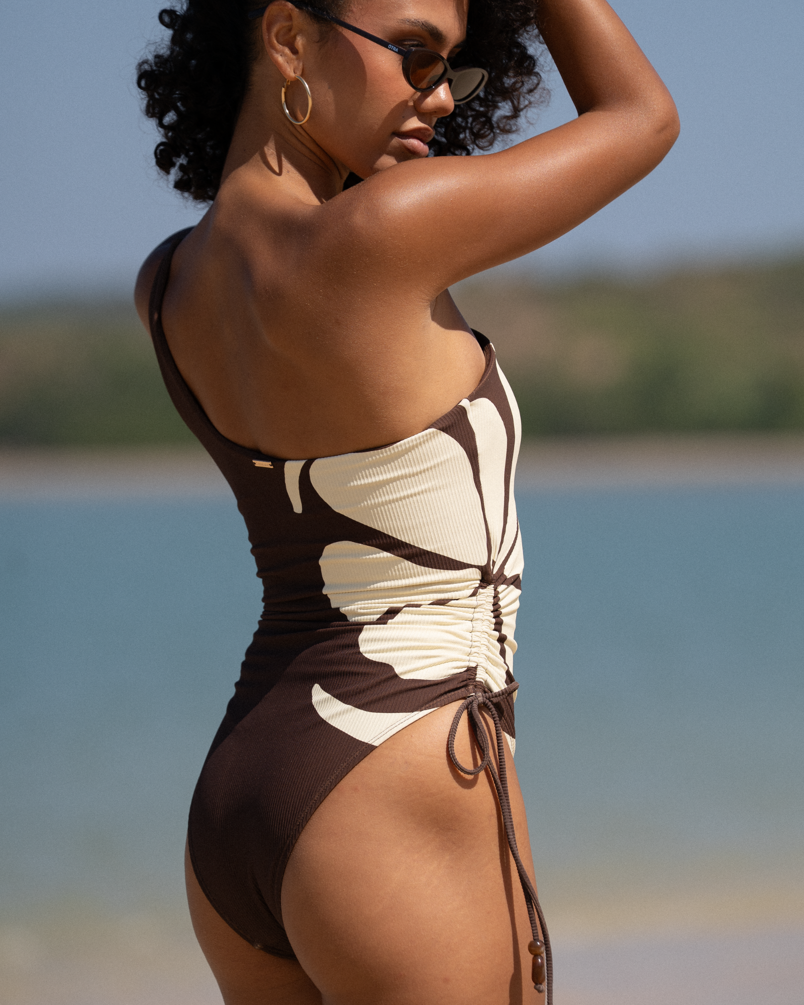Side view of chocolate brown ribbed one-shoulder swimsuit with cream floral panel and ruched tie detail, beach styled shot