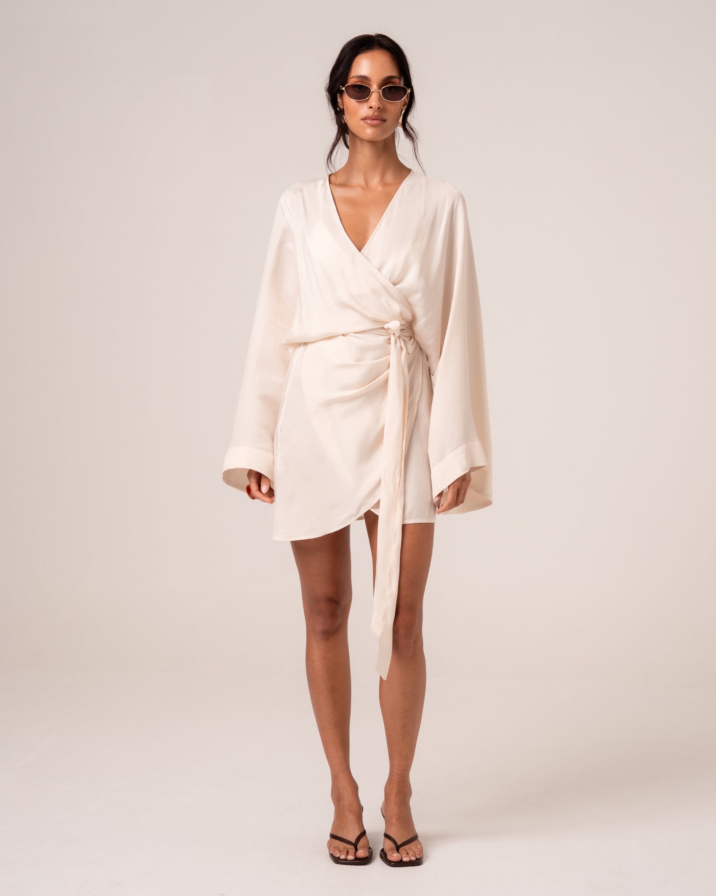 Model wearing an ivory batwing wrap mini dress with a deep V-neckline, wide sleeves and side tie, studio front view