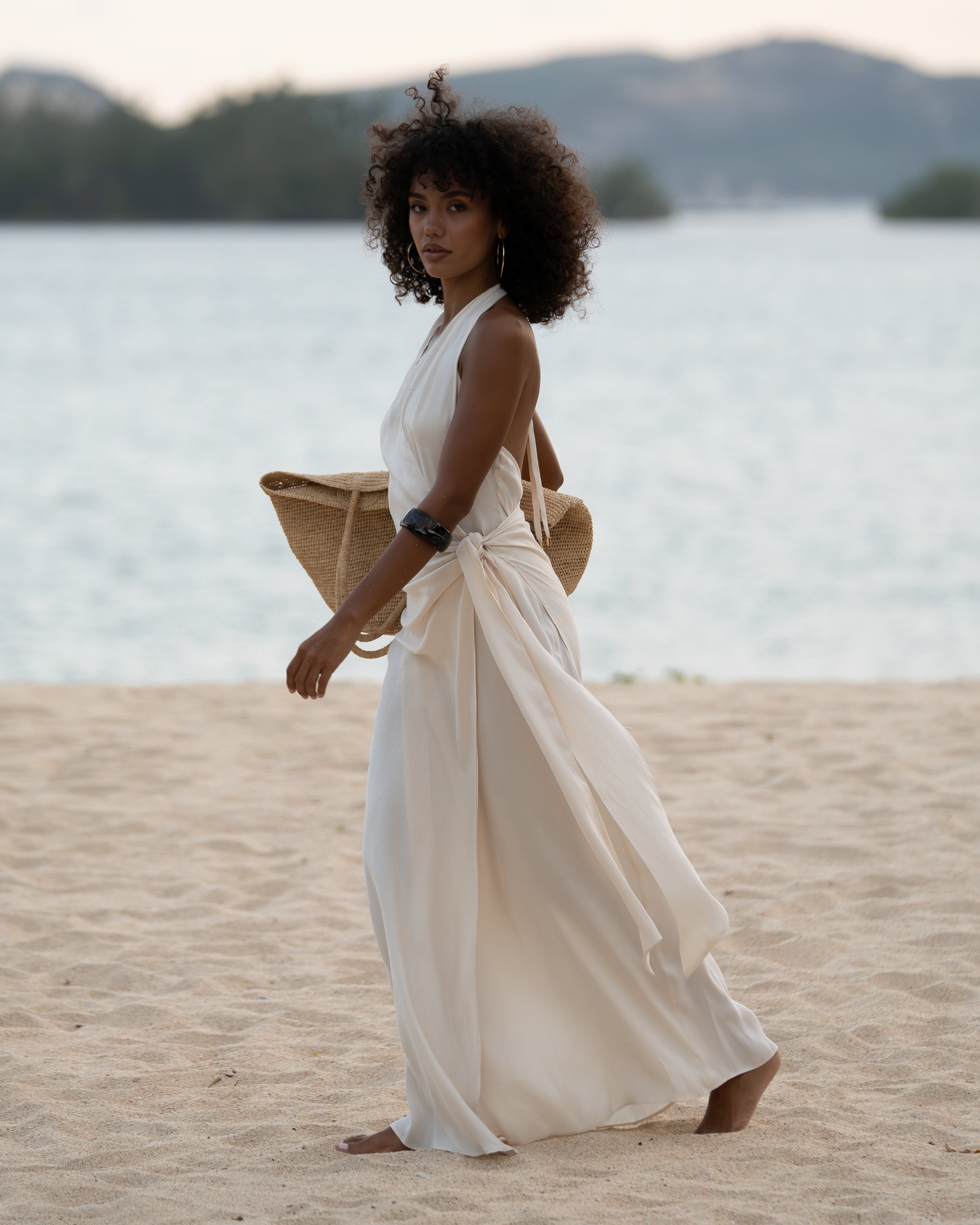 Woman walking on the beach in an ivory halter wrap maxi dress with cascading side tie, carrying a straw tote