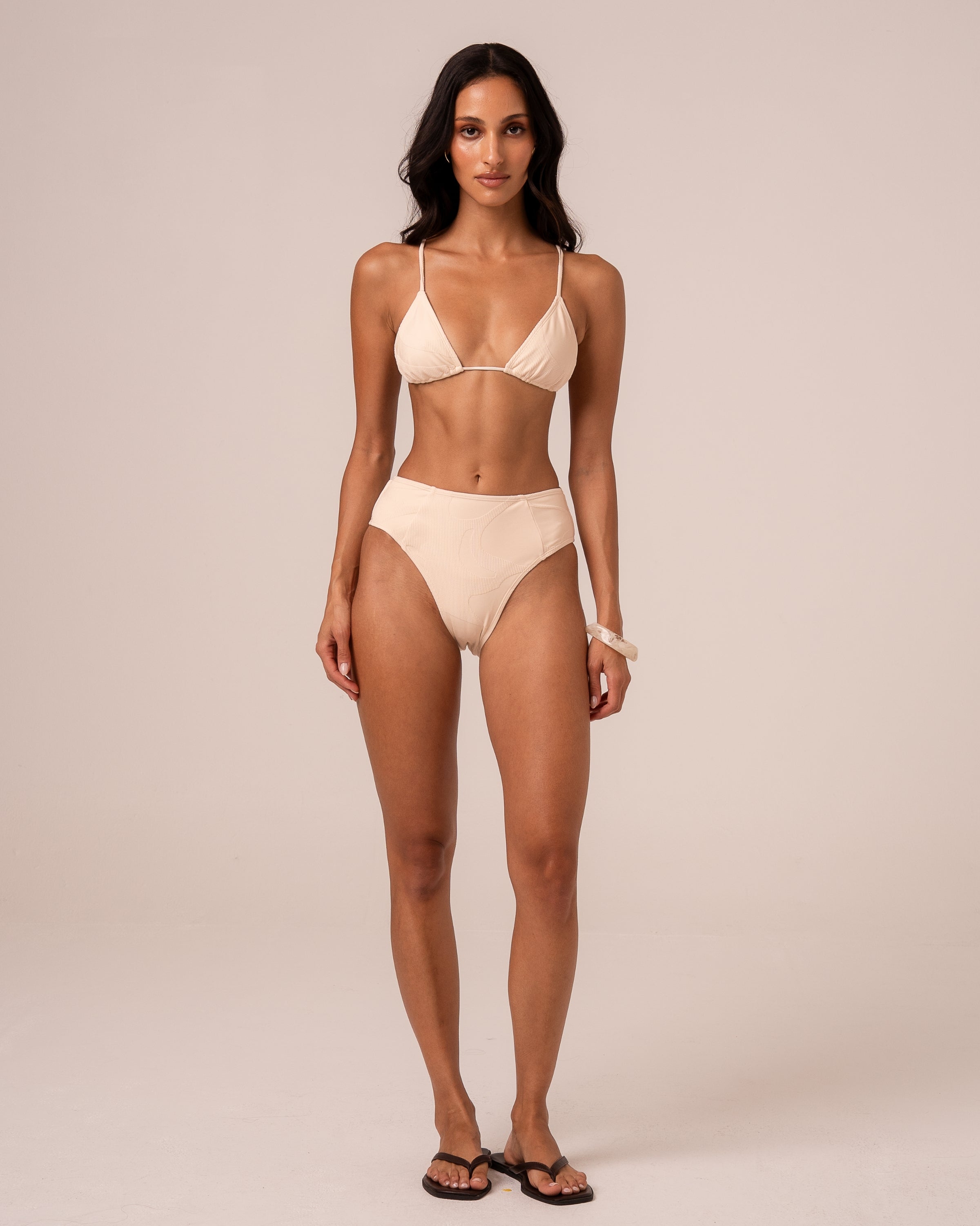 Model wearing an ivory mid rise bikini bottom with spliced panel seams and a higher leg, styled with a triangle top — studio front view