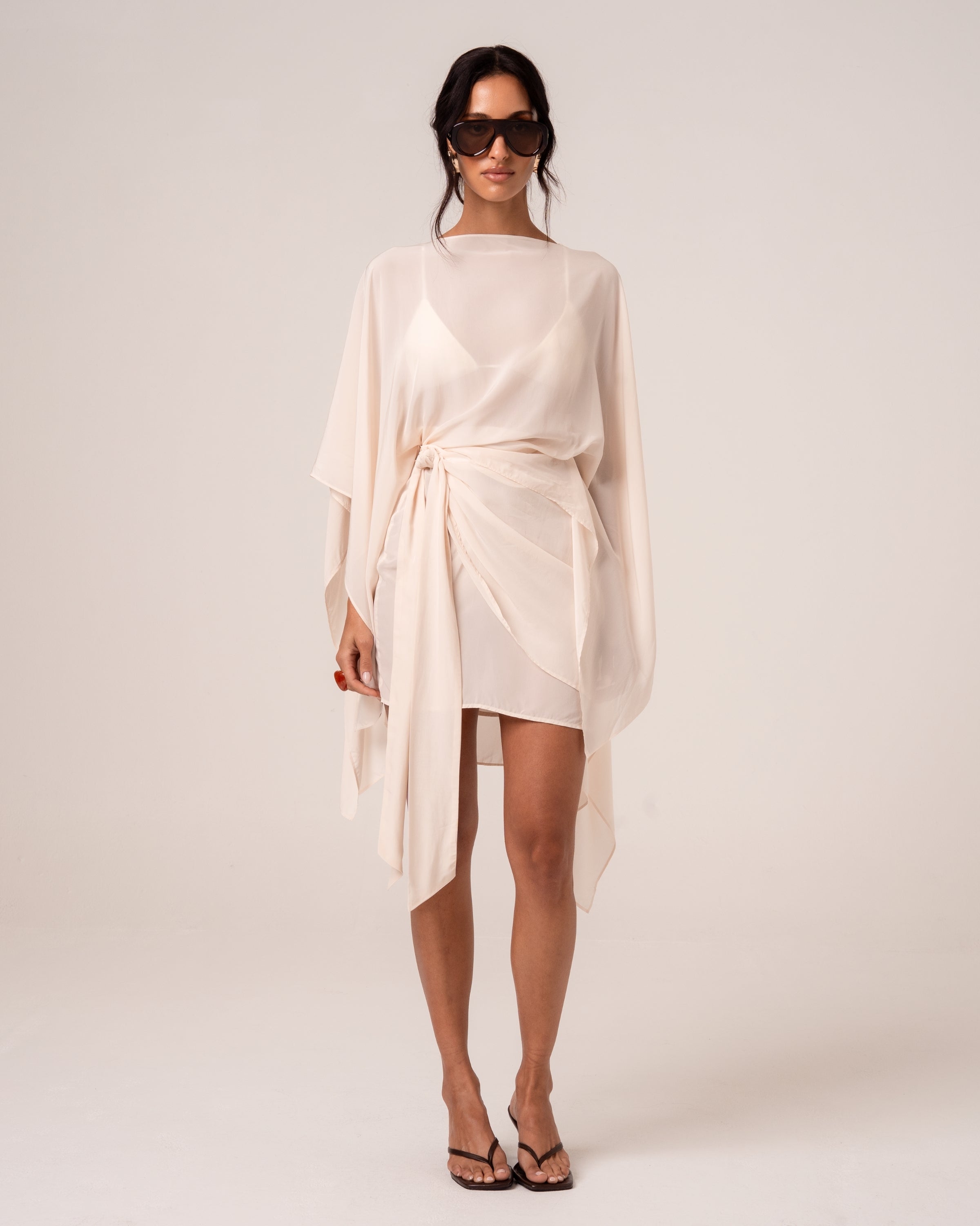 Front view of an ivory viscose wrap mini skirt with hip tie, styled with a sheer cape over a matching bikini