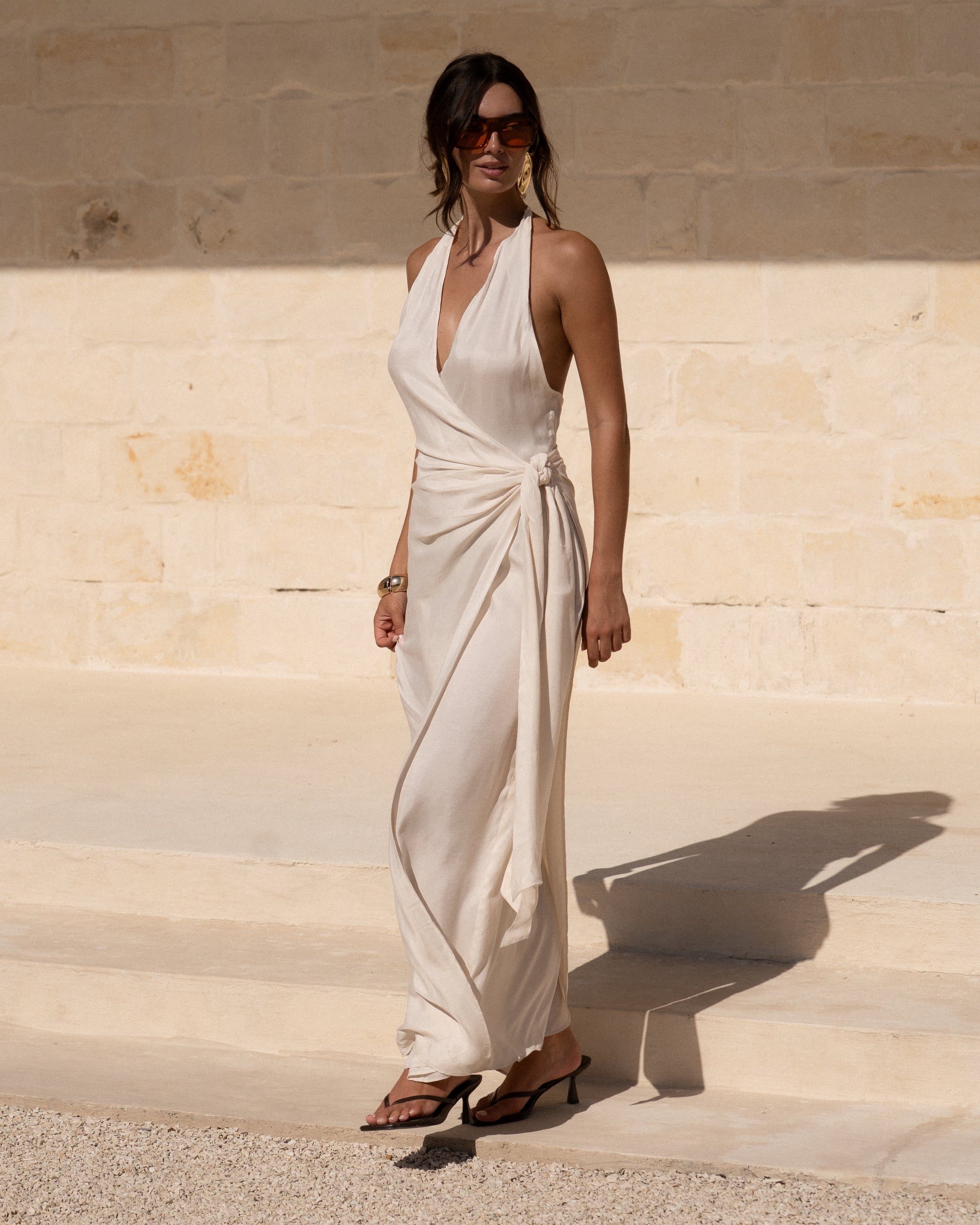 Ivory halter wrap maxi dress with plunging V-neckline and side knot worn with heels against a limestone wall — wedding guest resortwear