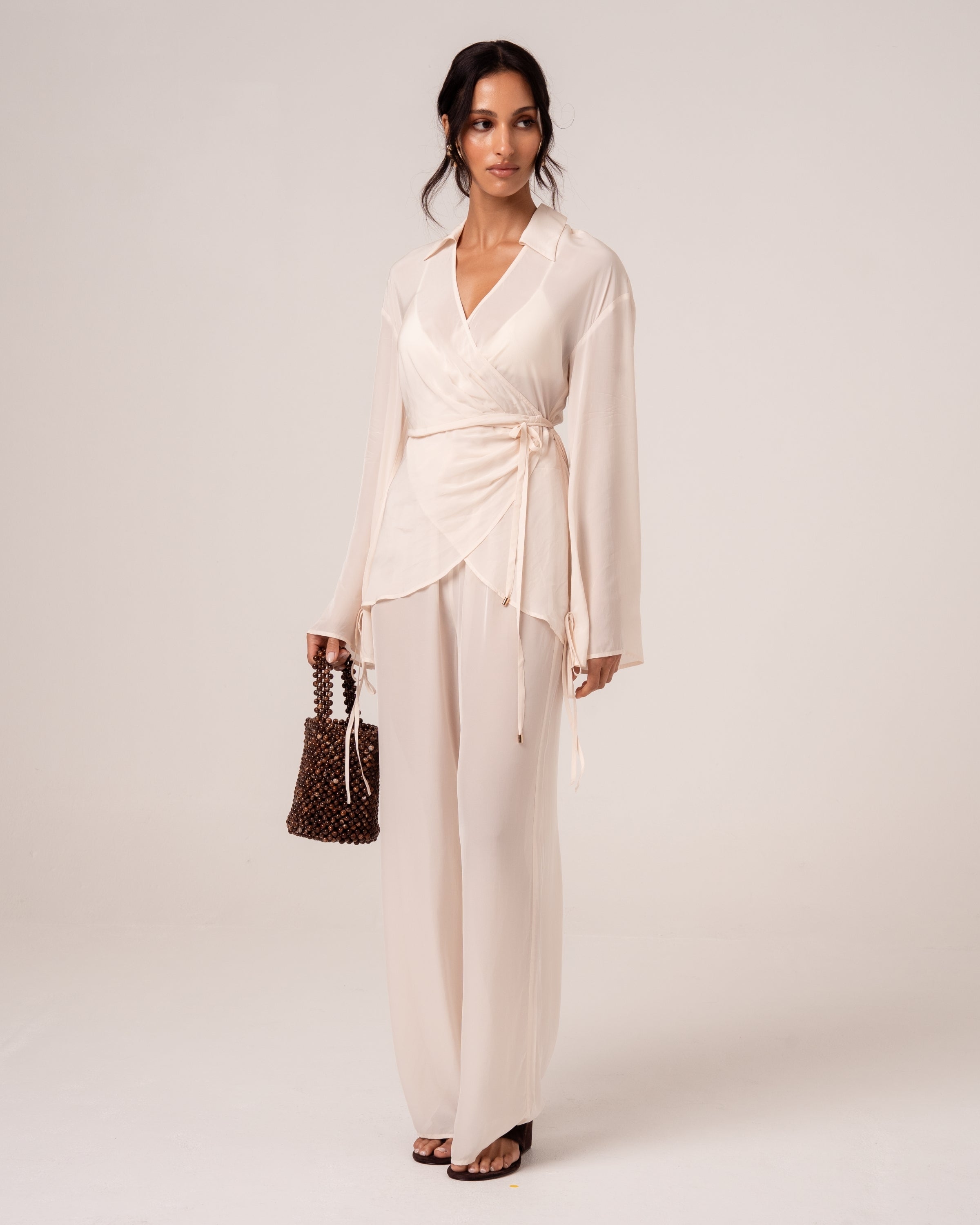 Side view of an ivory wrap shirt with waist tie worn with flowy wide-leg pants and a beaded bag
