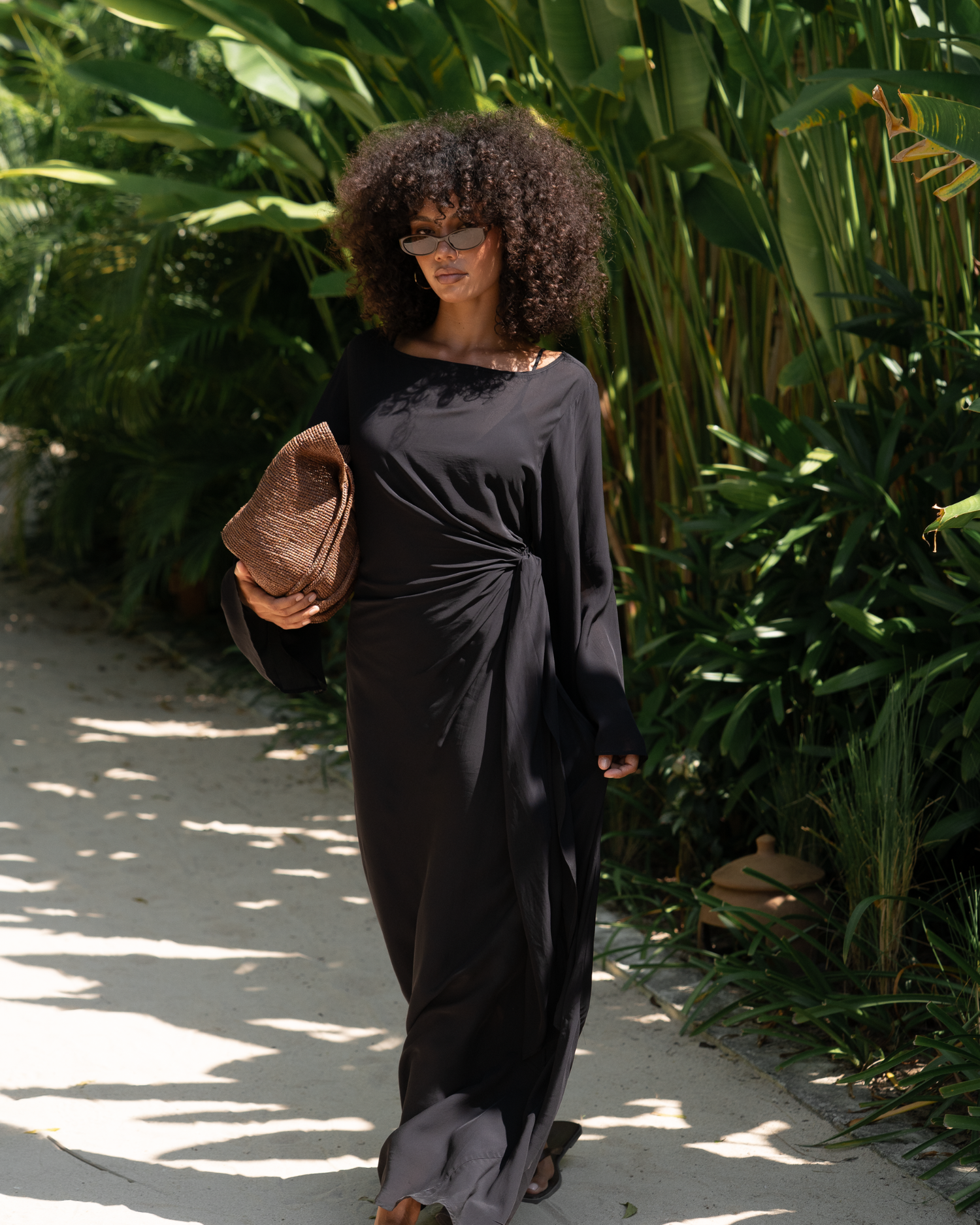 Woman in a black batwing maxi dress walking a garden path, wide sleeves and gathered waist knot, holding a raffia clutch