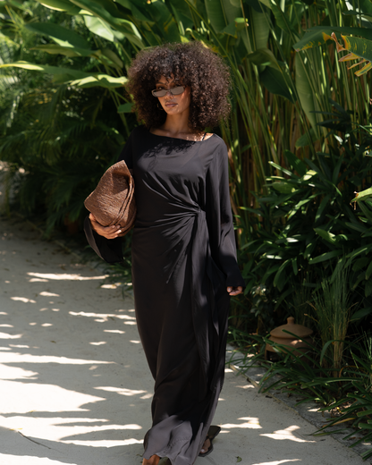 Woman in a black batwing maxi dress walking a garden path, wide sleeves and gathered waist knot, holding a raffia clutch