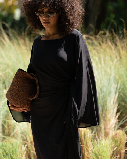 Close crop of black batwing maxi dress with gathered knot waist detail, styled in a grassy resort setting with a raffia clutch