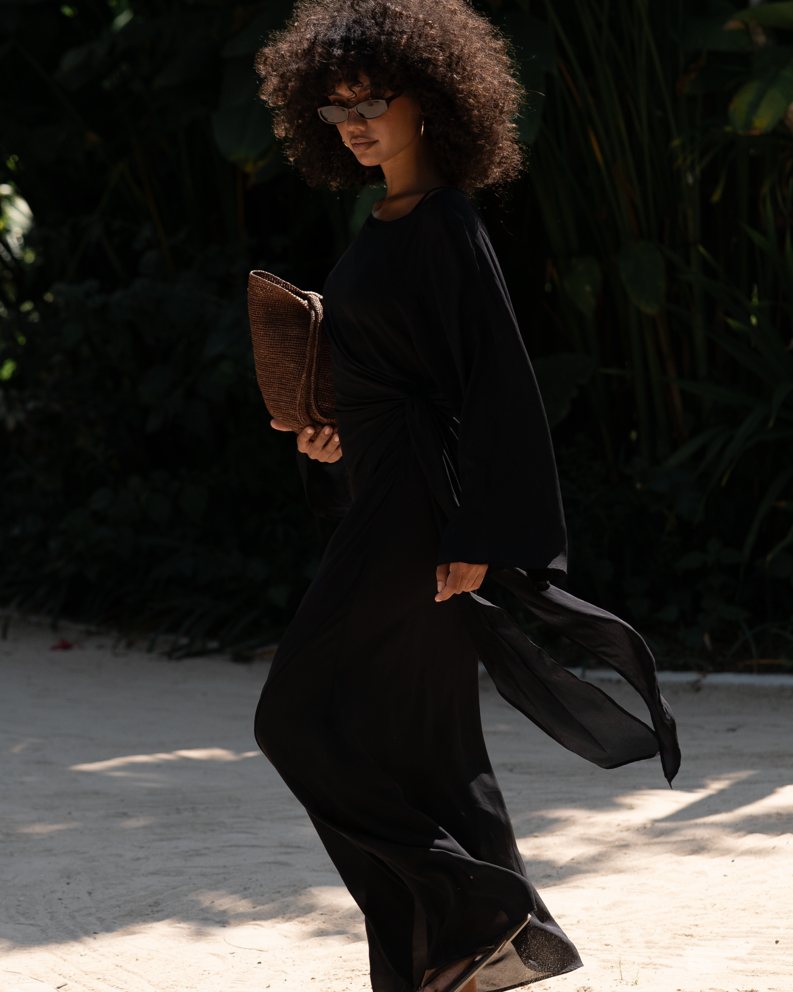 Side view of a black batwing maxi dress in motion, wide sleeve fabric flowing on a sandy resort path