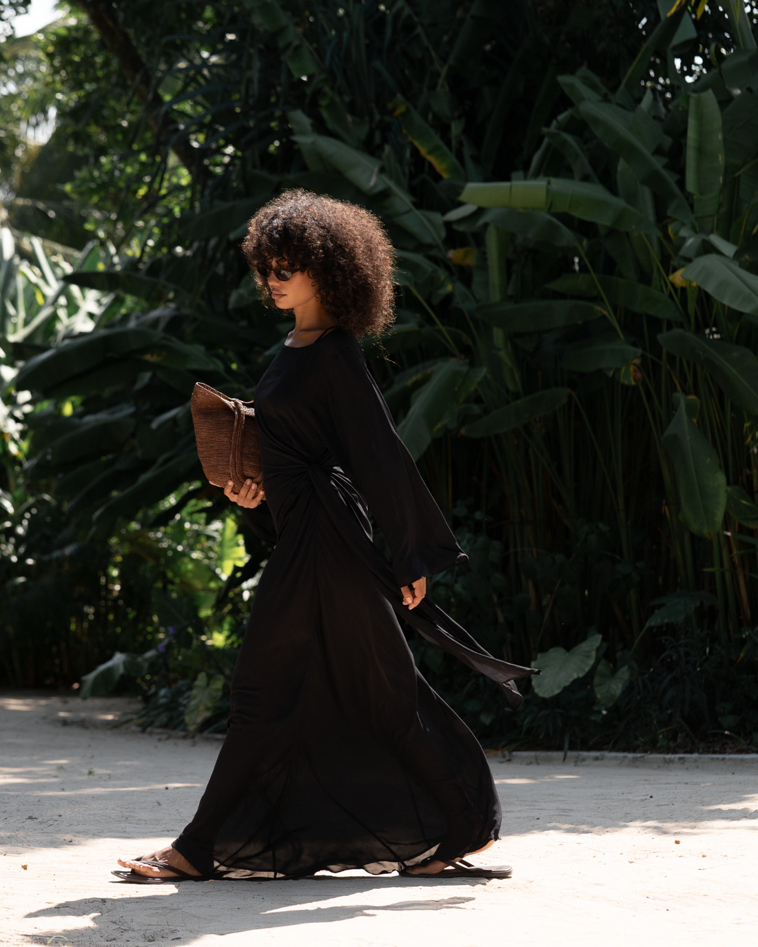 Woman walking through a tropical garden in a black batwing maxi dress with draped side knot, carrying a woven raffia clutch