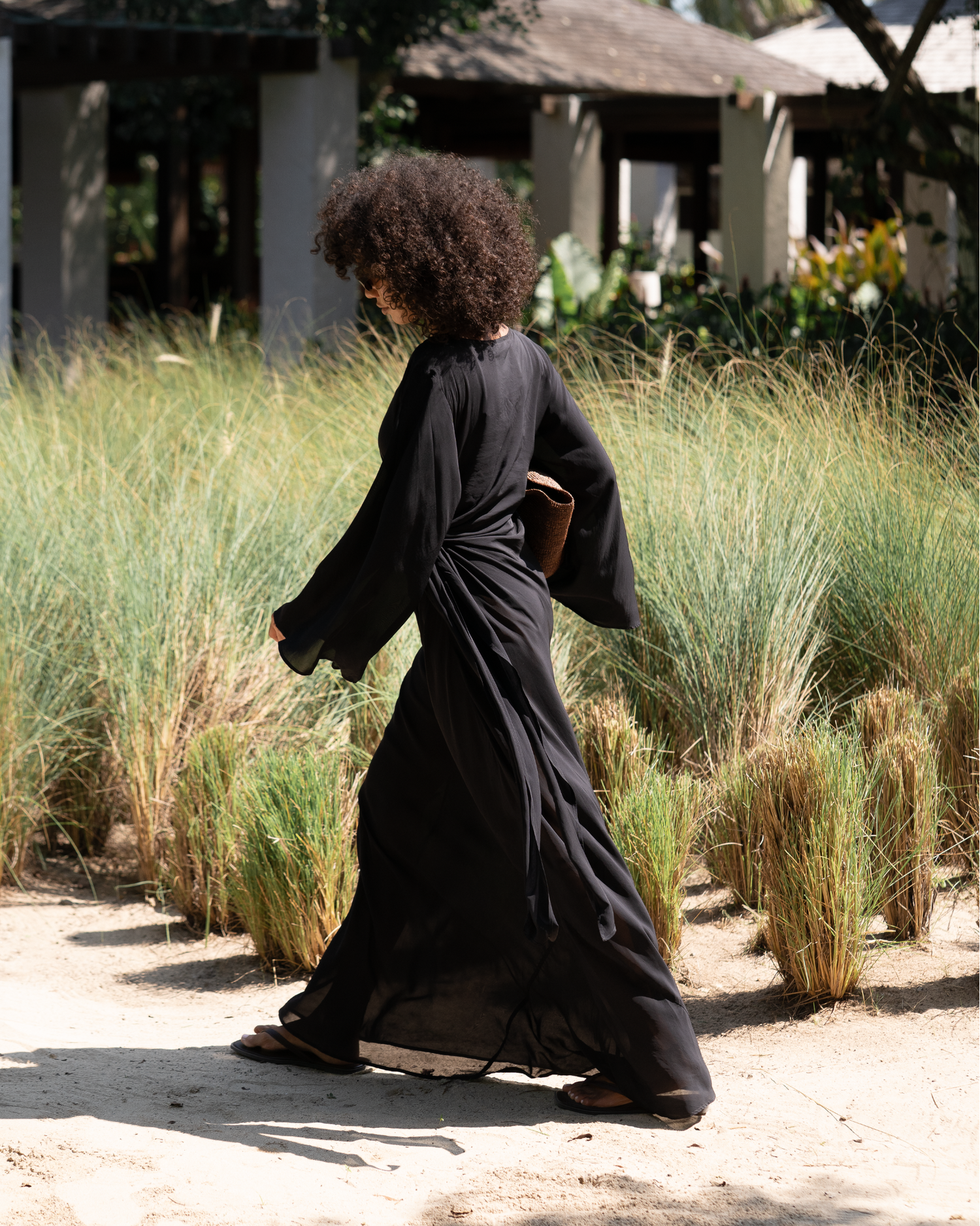 Back view of woman walking in a black batwing maxi dress past tall grasses, showing the flowing wide sleeves