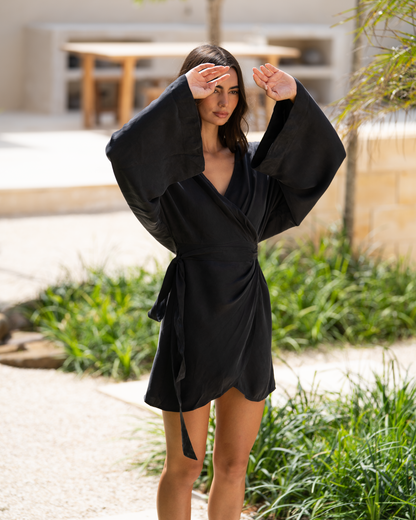 Front view of a model wearing a black batwing sleeve wrap mini dress outdoors, shading her eyes with dramatic flared sleeves visible