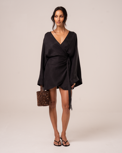 Front view of a model wearing a black batwing sleeve wrap mini dress with a deep V neckline, styled with a beaded bag