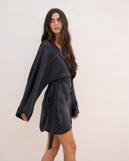Side view of a model walking in a black batwing sleeve wrap mini dress against a plain studio wall