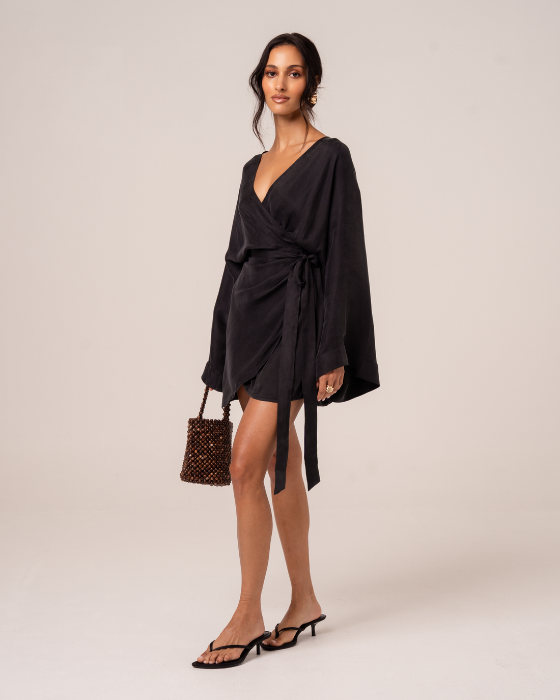 Side view of a model wearing a black batwing sleeve wrap mini dress with a draped waist tie, studio side view