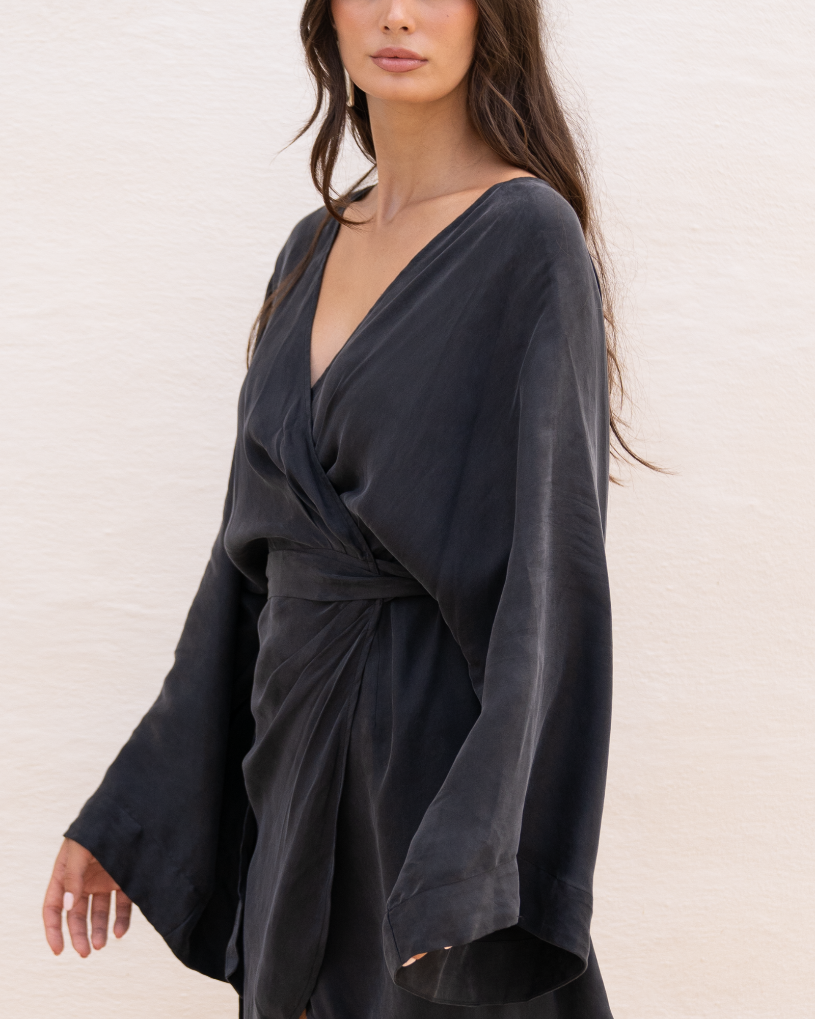 Close-up of a model wearing a black batwing sleeve wrap mini dress showing the deep V neckline and knotted waist tie
