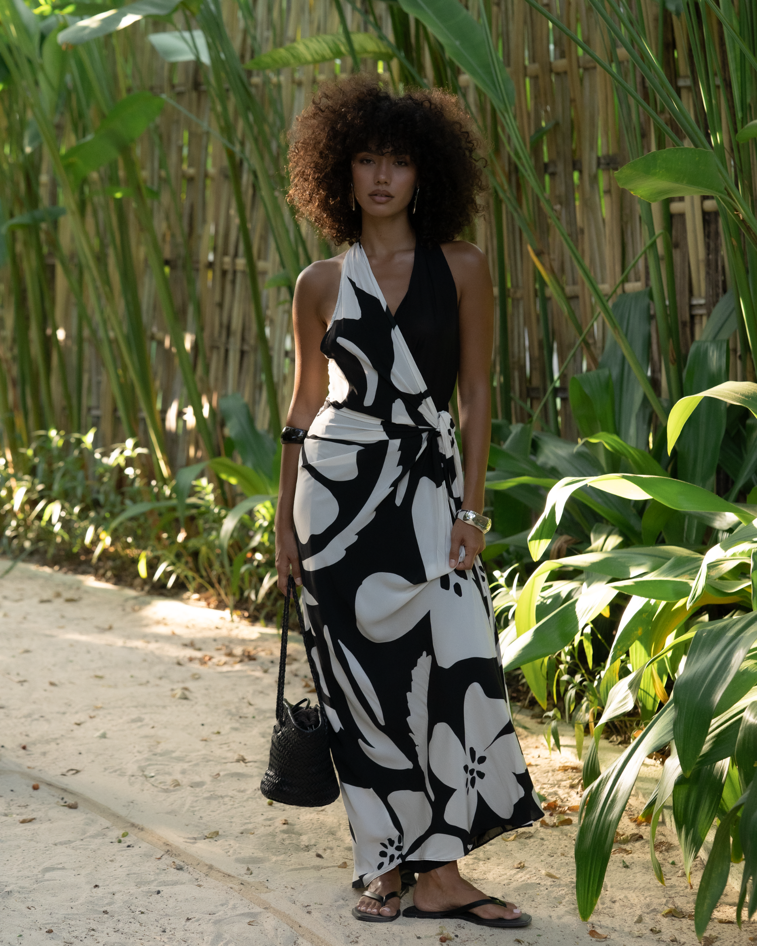 Front view of a model wearing a black and cream floral halter wrap maxi dress standing among bamboo, styled with a woven bag