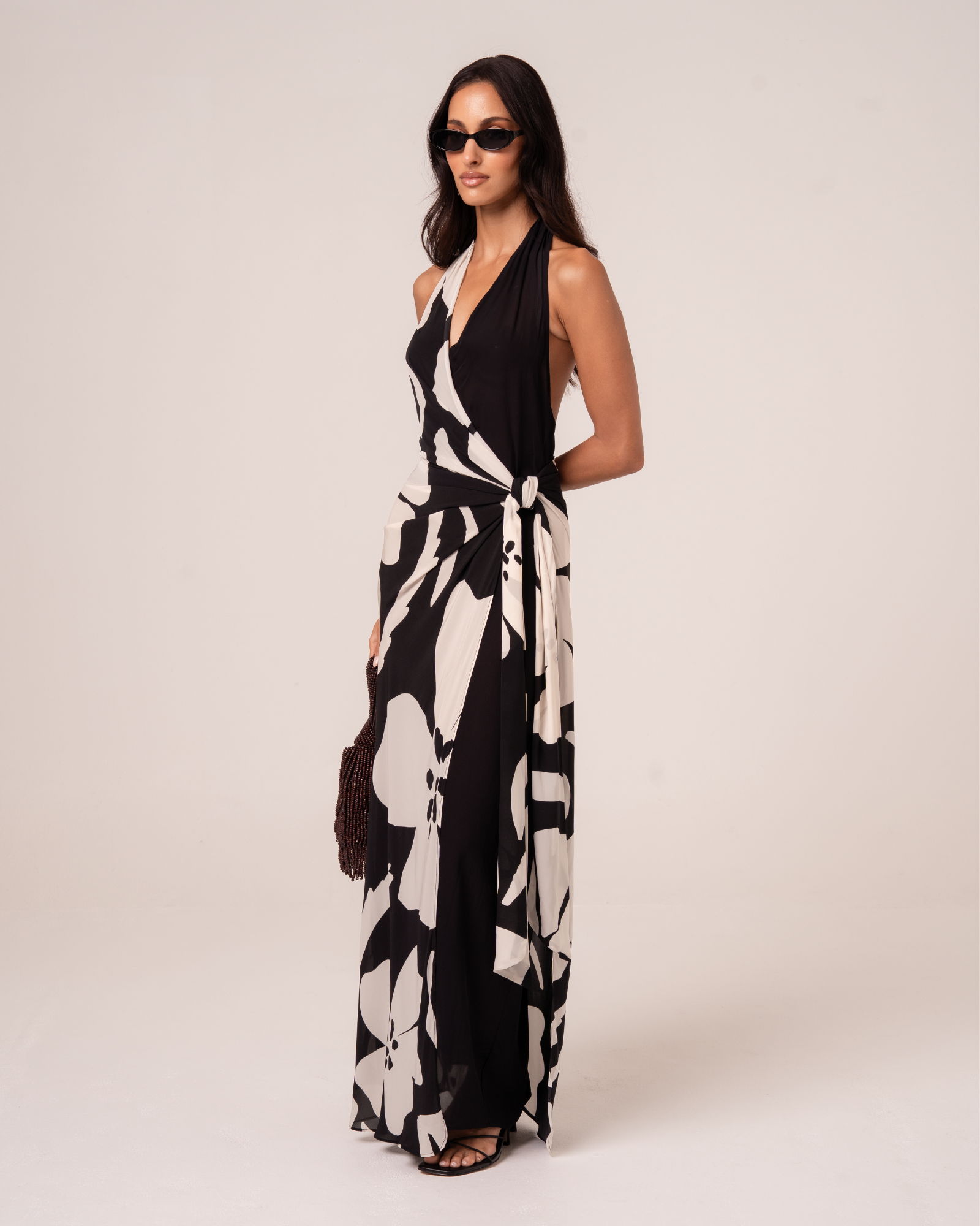 Side view of a model wearing a black and cream floral halter wrap maxi dress with a draped sash tie waist, studio side view