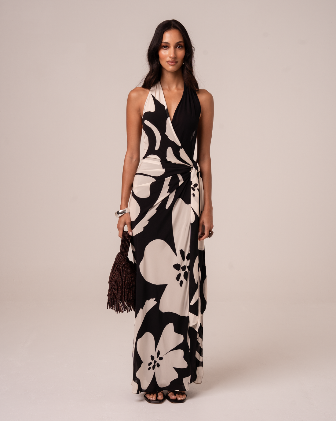 Front view of a model wearing a black and cream floral halter wrap maxi dress with a plunging neckline and draped tie waist, studio front view