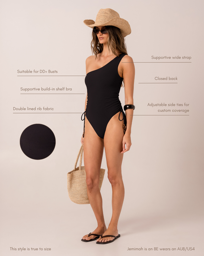 Fit and feature diagram of a black ribbed one-shoulder one-piece swimsuit showing supportive wide strap, closed back, adjustable side ties, and built-in shelf bra for DD+ busts