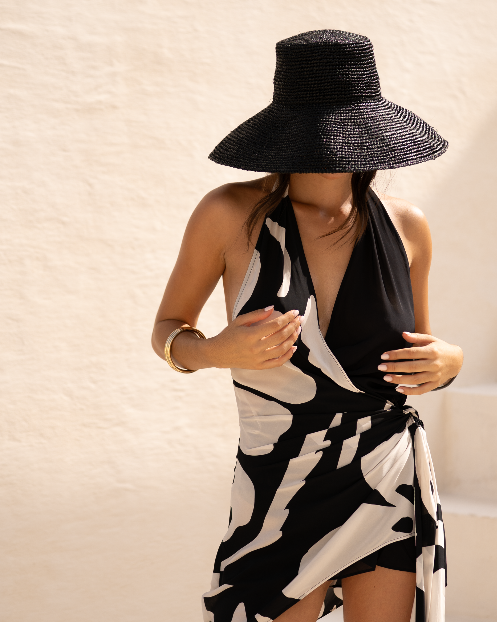 Black raffia straw bucket hat with a wide brim styled with a monochrome print halter dress against a whitewashed wall