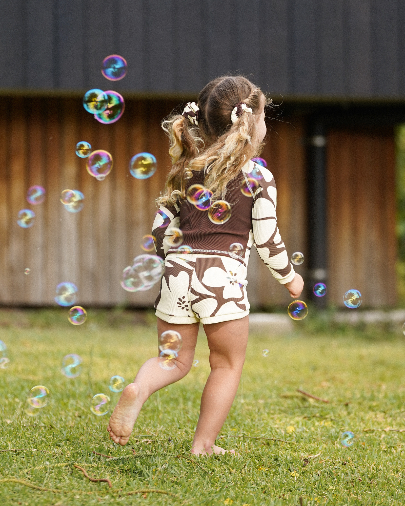 Girl standing amid floating bubbles in a brown and cream hibiscus floral print long sleeve rash shirt and matching shorts, back view