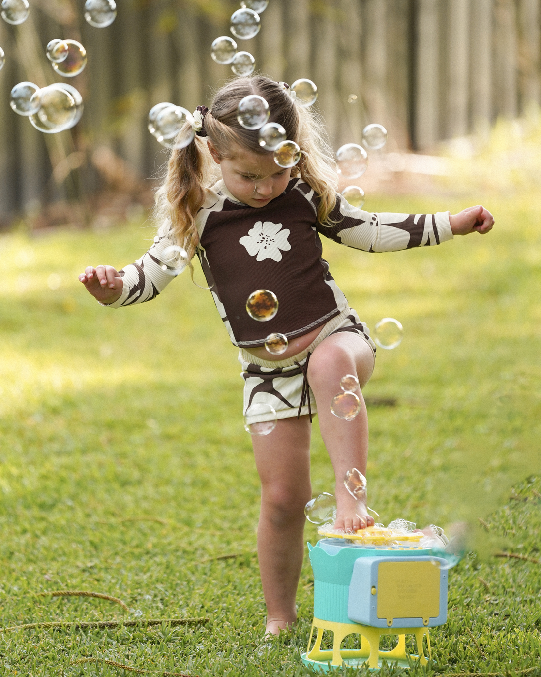 Girl playing with a bubble machine outdoors wearing a brown and cream hibiscus floral print long sleeve rash shirt and matching shorts