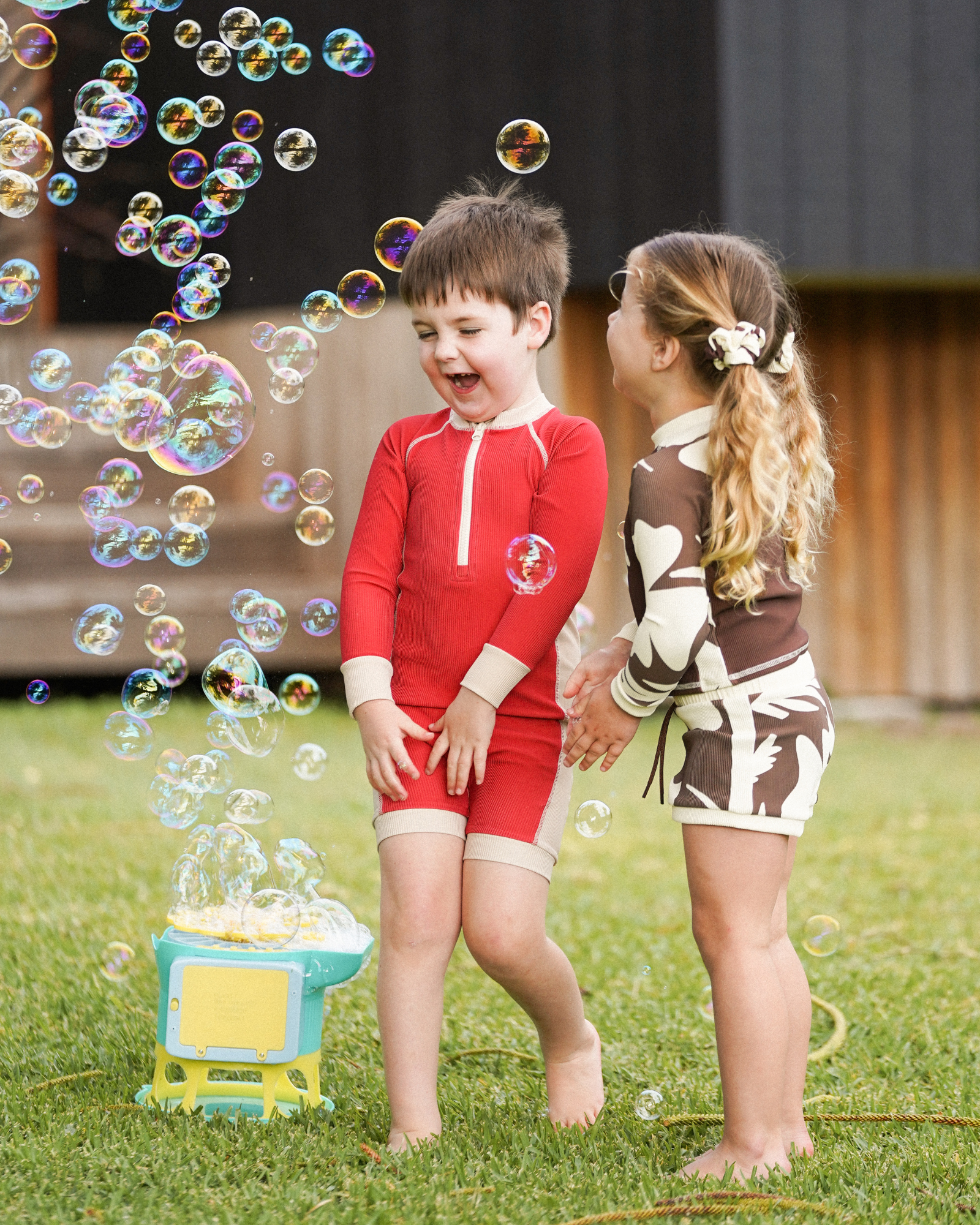 Brown and cream hibiscus print kids swim shorts with matching rash top worn by girl playing with a boy in a red swim outfit, front view lifestyle