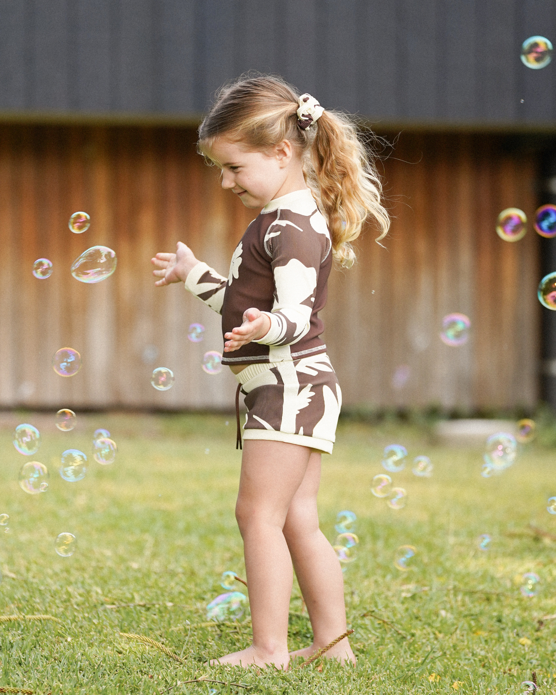Brown and cream hibiscus print kids swim shorts with matching rash top, side view, girl playing with bubbles outdoors