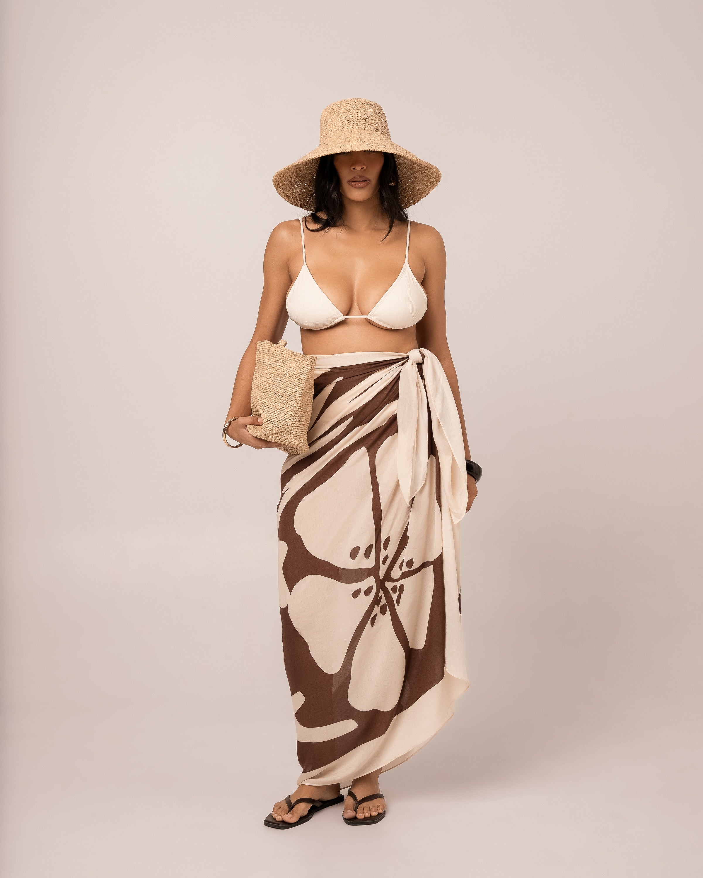 Ochre Lane Ibisco Sarong in brown hibiscus print worn as a beach cover-up with straw hat and woven basket bag