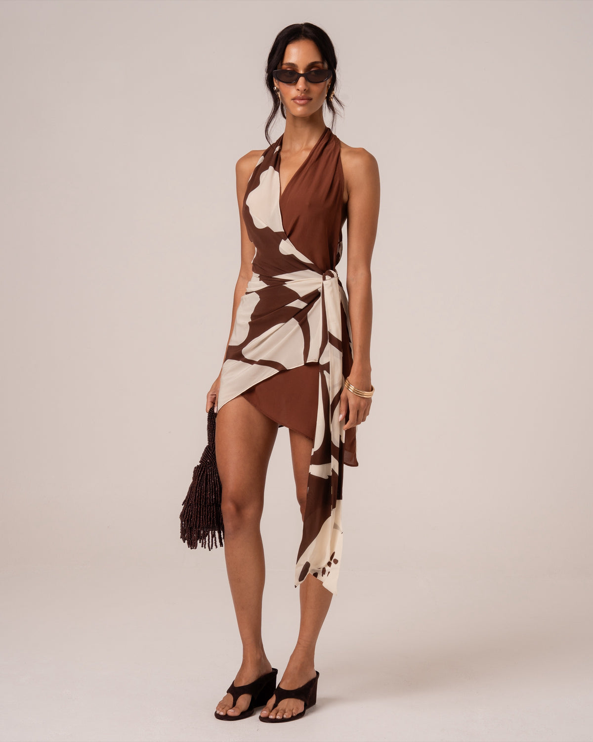 Front view of a brown and cream floral wrap mini dress showing the draped side tie and asymmetric hem, styled with a beaded bag