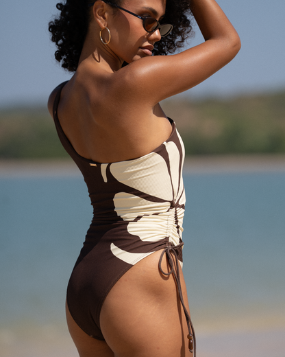 Side view of chocolate brown ribbed one-shoulder swimsuit with cream floral panel and ruched tie detail, beach styled shot