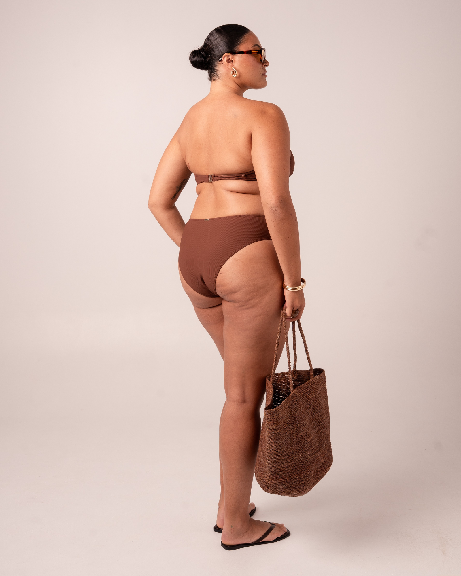Back view of a chocolate-brown ribbed splice mid-rise bikini bottom with a matching strapless bandeau top, carrying a woven tote bag