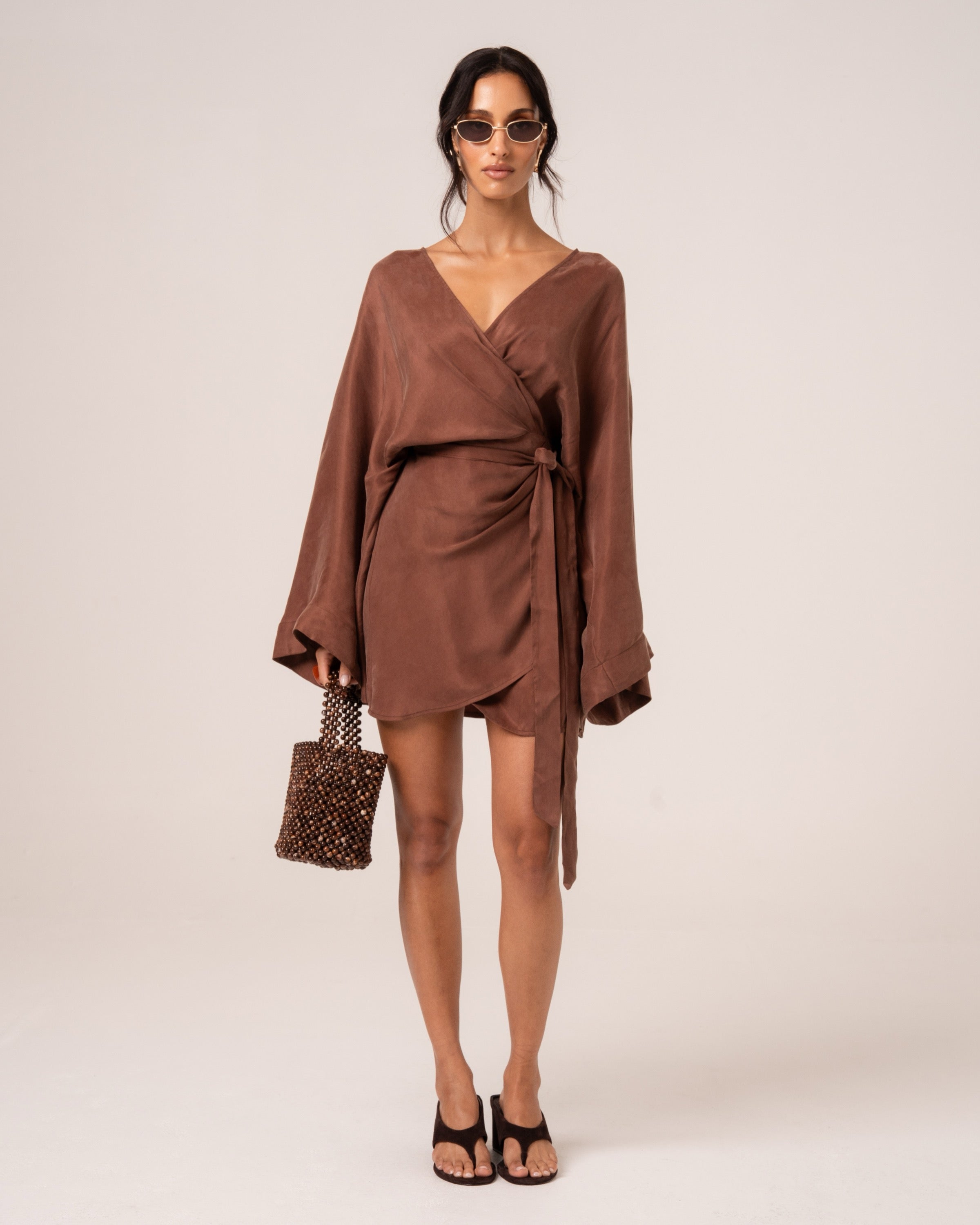 Front view of a chocolate brown batwing wrap mini dress with a deep V-neck and side waist tie, styled with a beaded bag