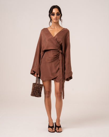 Front view of a chocolate brown batwing wrap mini dress with a deep V-neck and side waist tie, styled with a beaded bag
