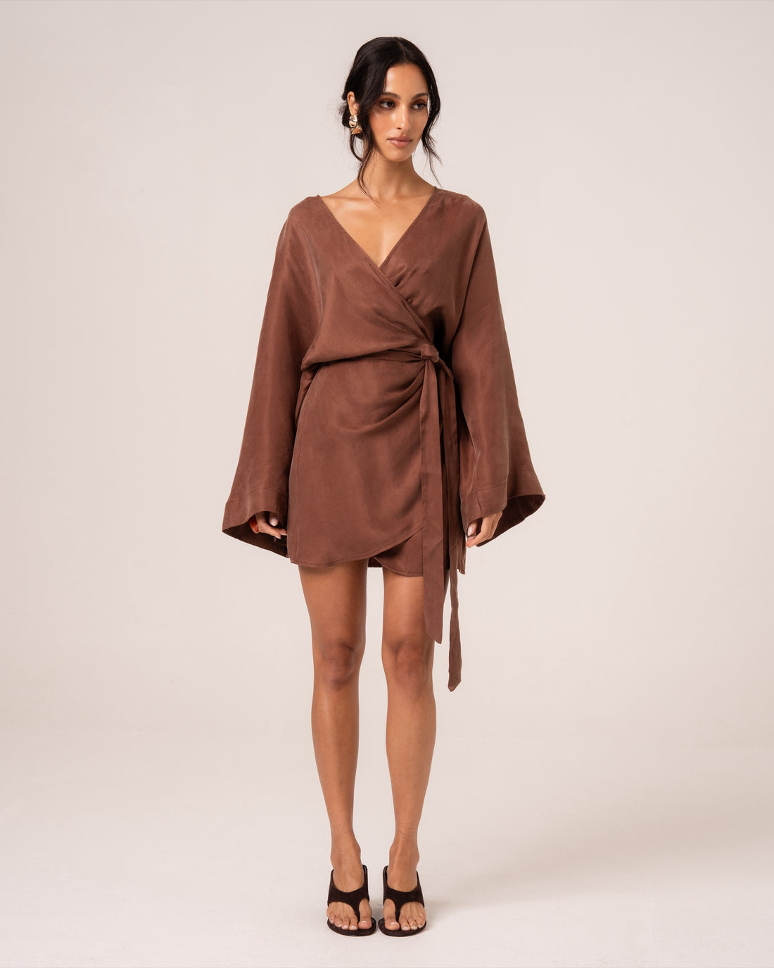 Model wearing a chocolate brown batwing wrap mini dress with a deep V-neckline and wide sleeves, studio front view