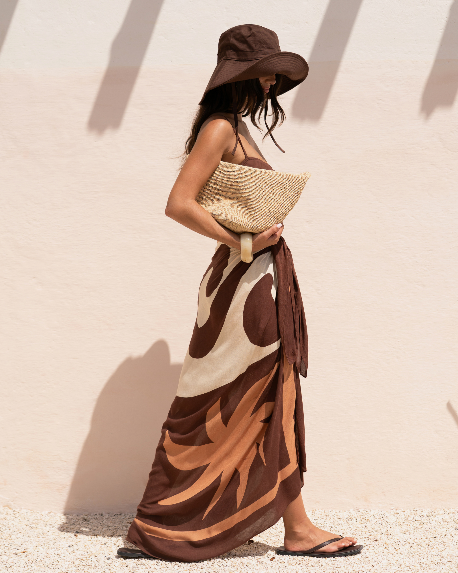 Model wearing a chocolate, cream, and tan abstract floral print sarong tied as a long wrap skirt, side view walking against a sunlit wall, styled with a brown bucket hat and raffia clutch
