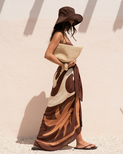 Model wearing a chocolate, cream, and tan abstract floral print sarong tied as a long wrap skirt, side view walking against a sunlit wall, styled with a brown bucket hat and raffia clutch