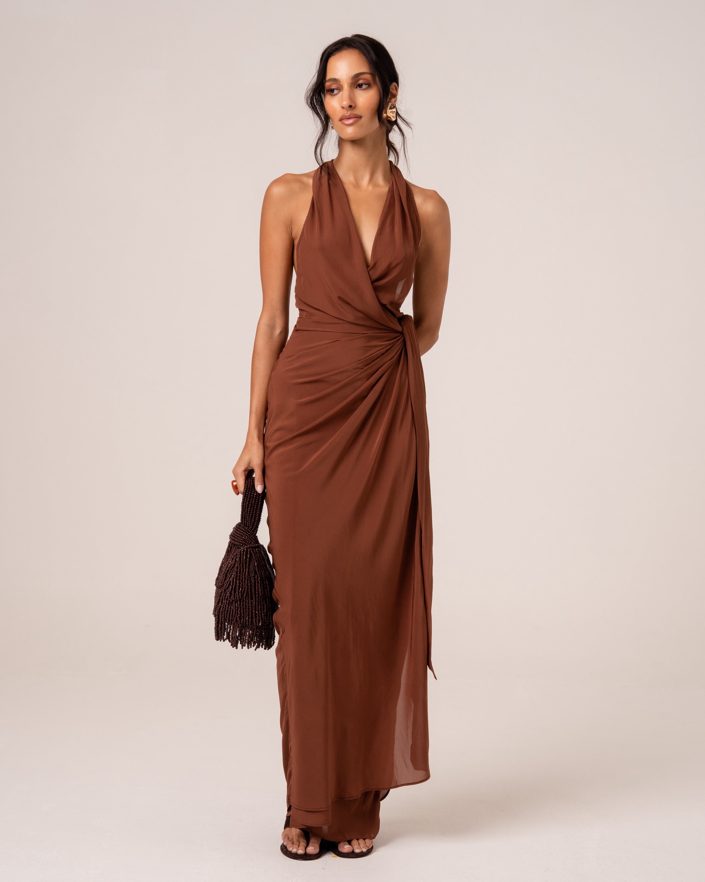 Model wearing a chocolate brown halter wrap maxi dress with a plunging neckline and knotted side tie, studio front view