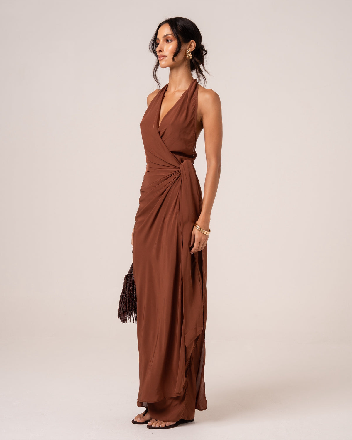 Side view of a chocolate brown halter wrap maxi dress showing the draped side knot and floor-length silhouette