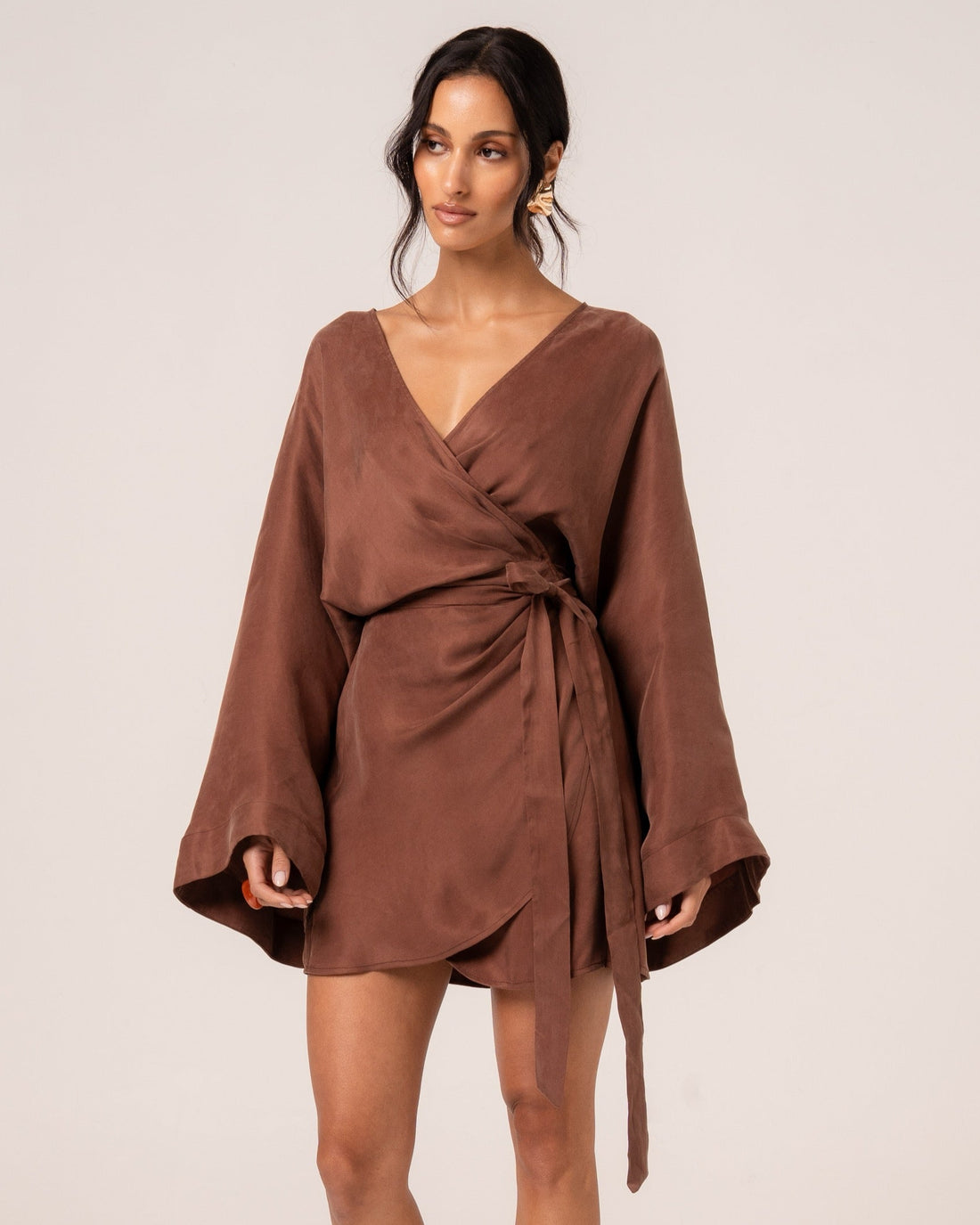 Front view of a chocolate brown batwing wrap mini dress showing the side tie waist and tulip hem, studio setting