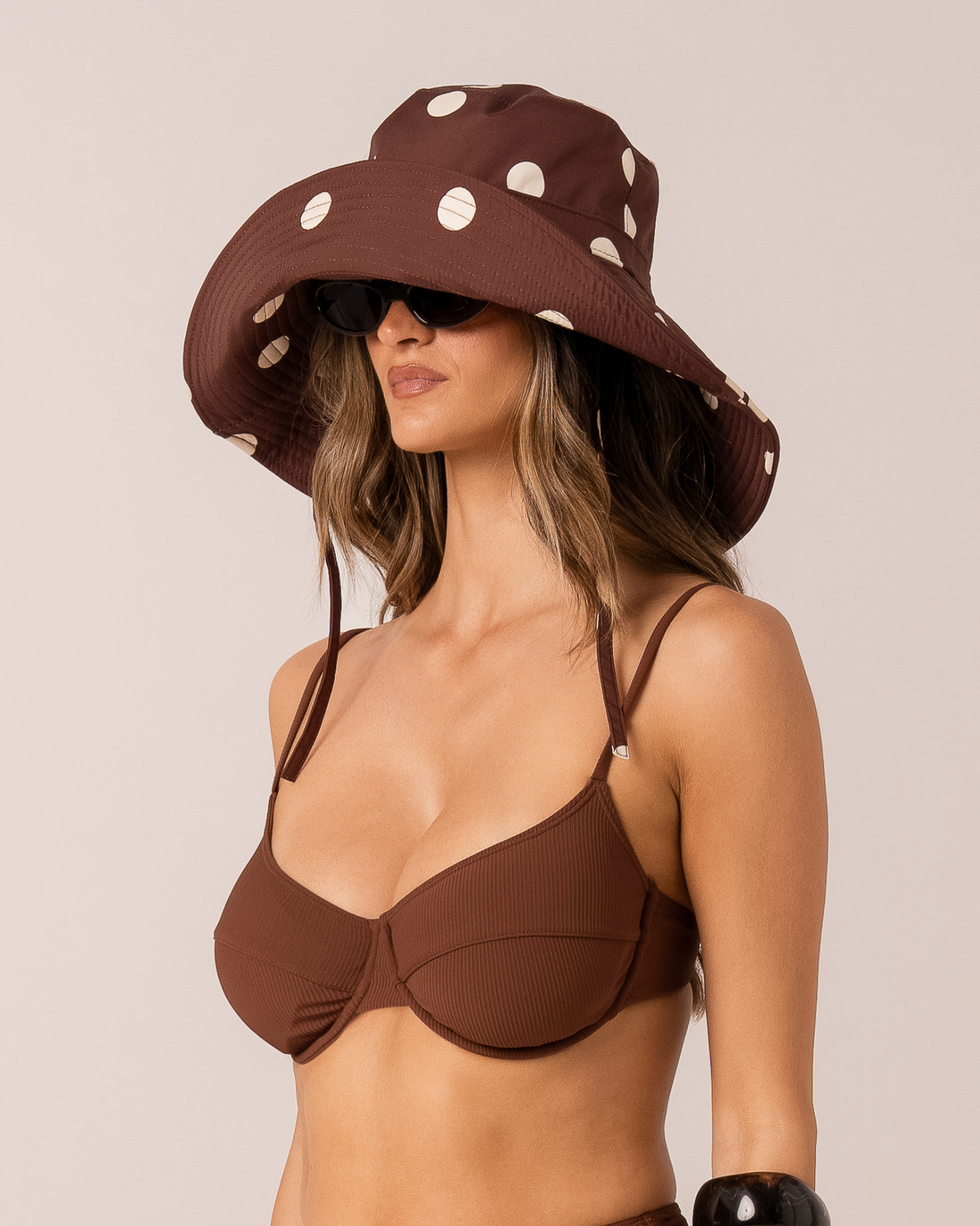 Model wearing a chocolate brown polka dot wide-brim sun hat, three-quarter view with sunglasses
