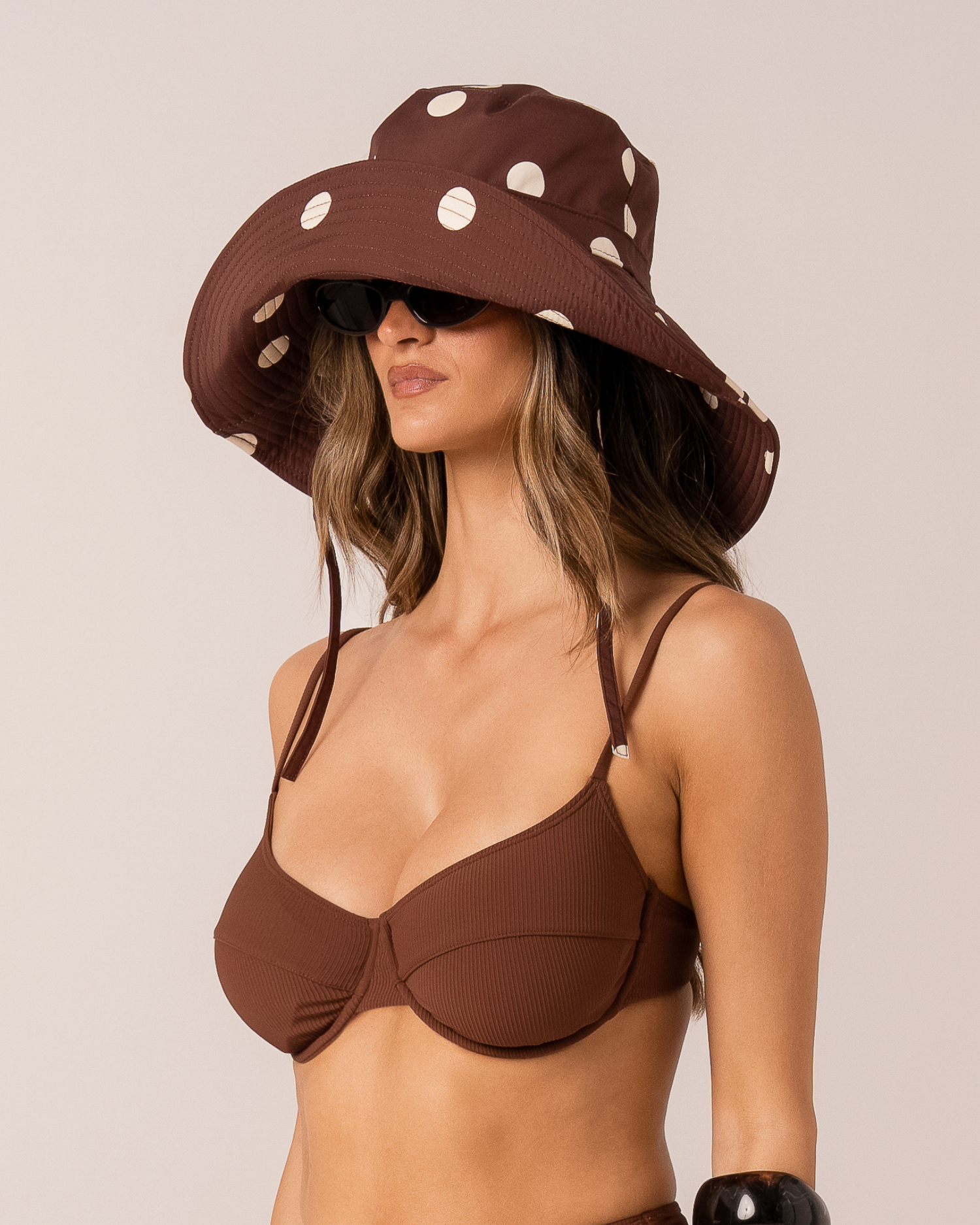 Model wearing a chocolate brown polka dot wide-brim sun hat, three-quarter view with sunglasses