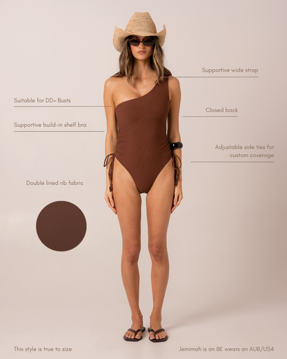 Fit and feature diagram of a chocolate ribbed one-shoulder one-piece swimsuit showing supportive wide strap, closed back, adjustable side ties, and built-in shelf bra for DD+ busts