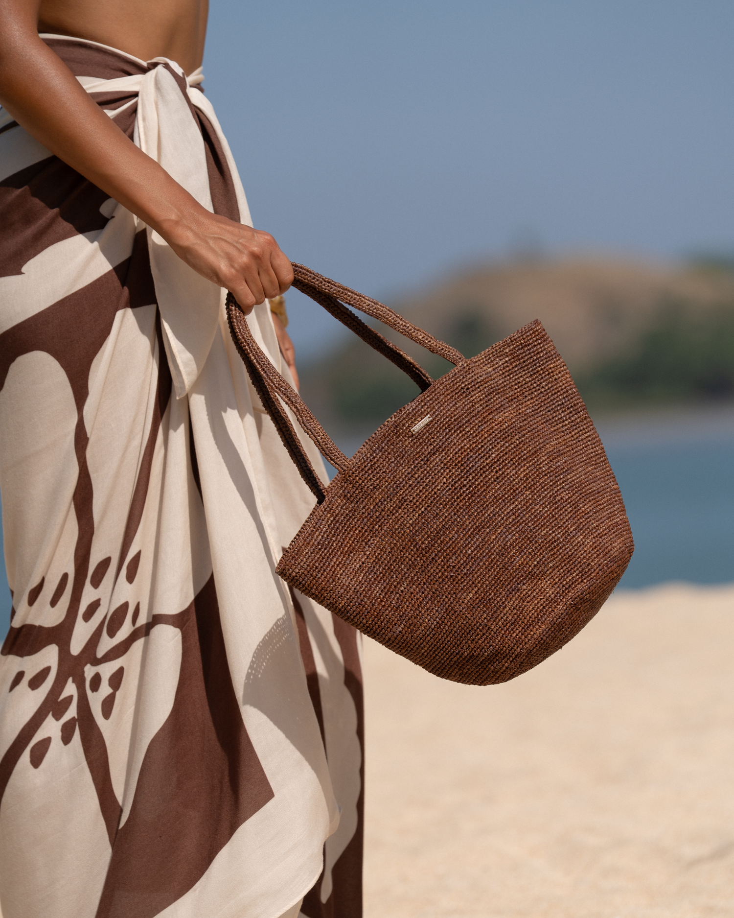 Close-up of a cocoa brown woven raffia mini tote held by the handle against a brown and cream printed sarong, beach background