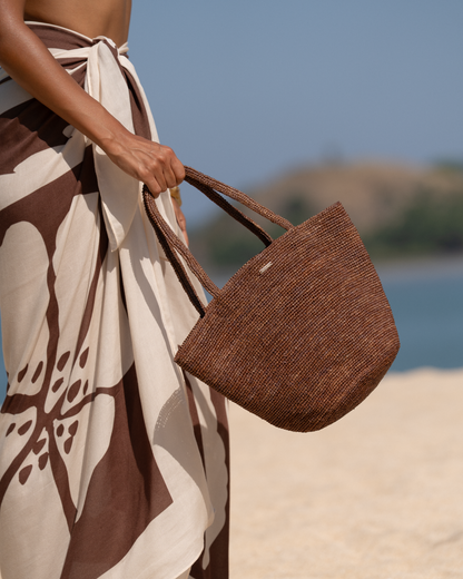 Close-up of a cocoa brown woven raffia mini tote held by the handle against a brown and cream printed sarong, beach background