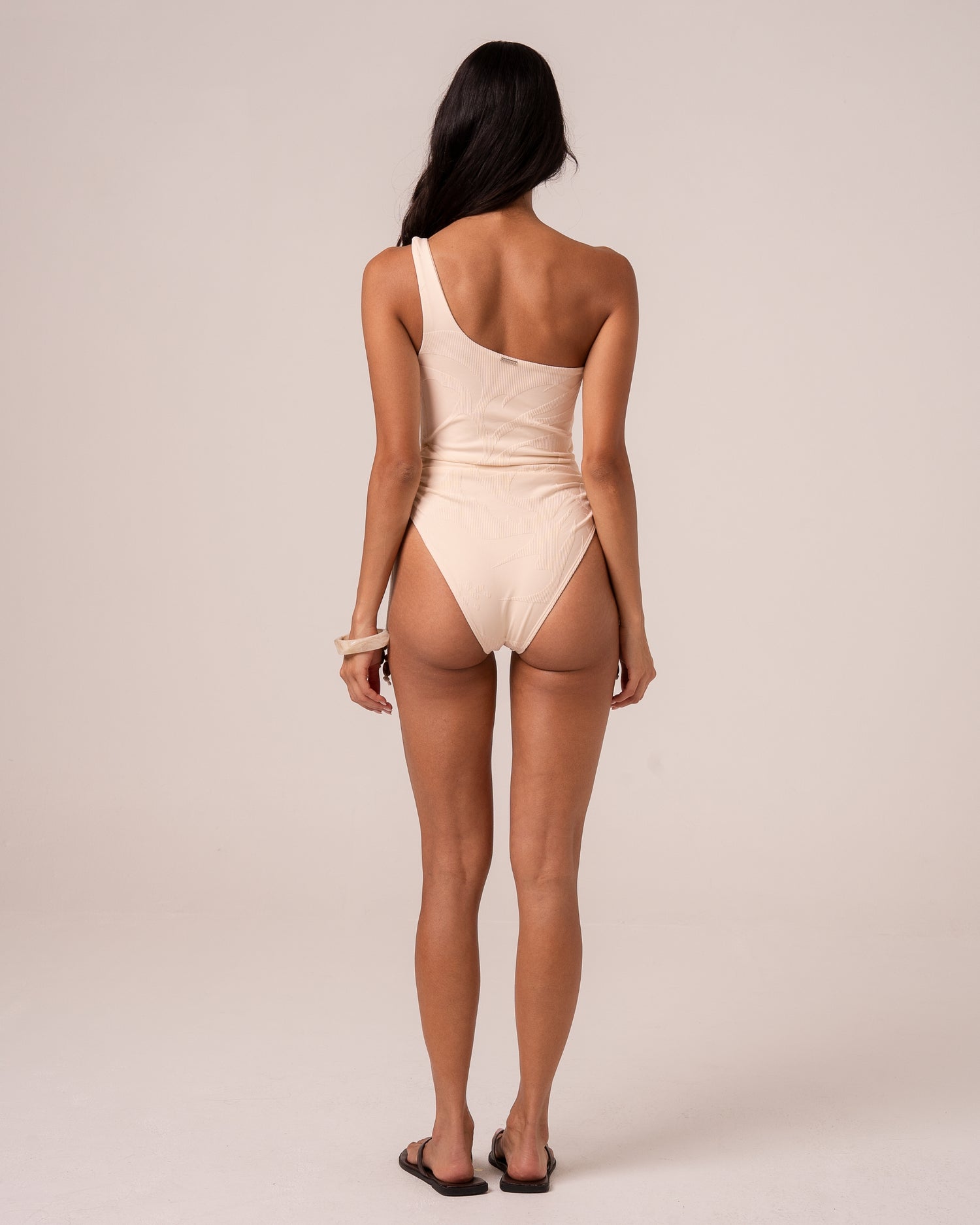 Back view of an ivory asym one shoulder one piece swimsuit showing the single shoulder strap and high-cut leg