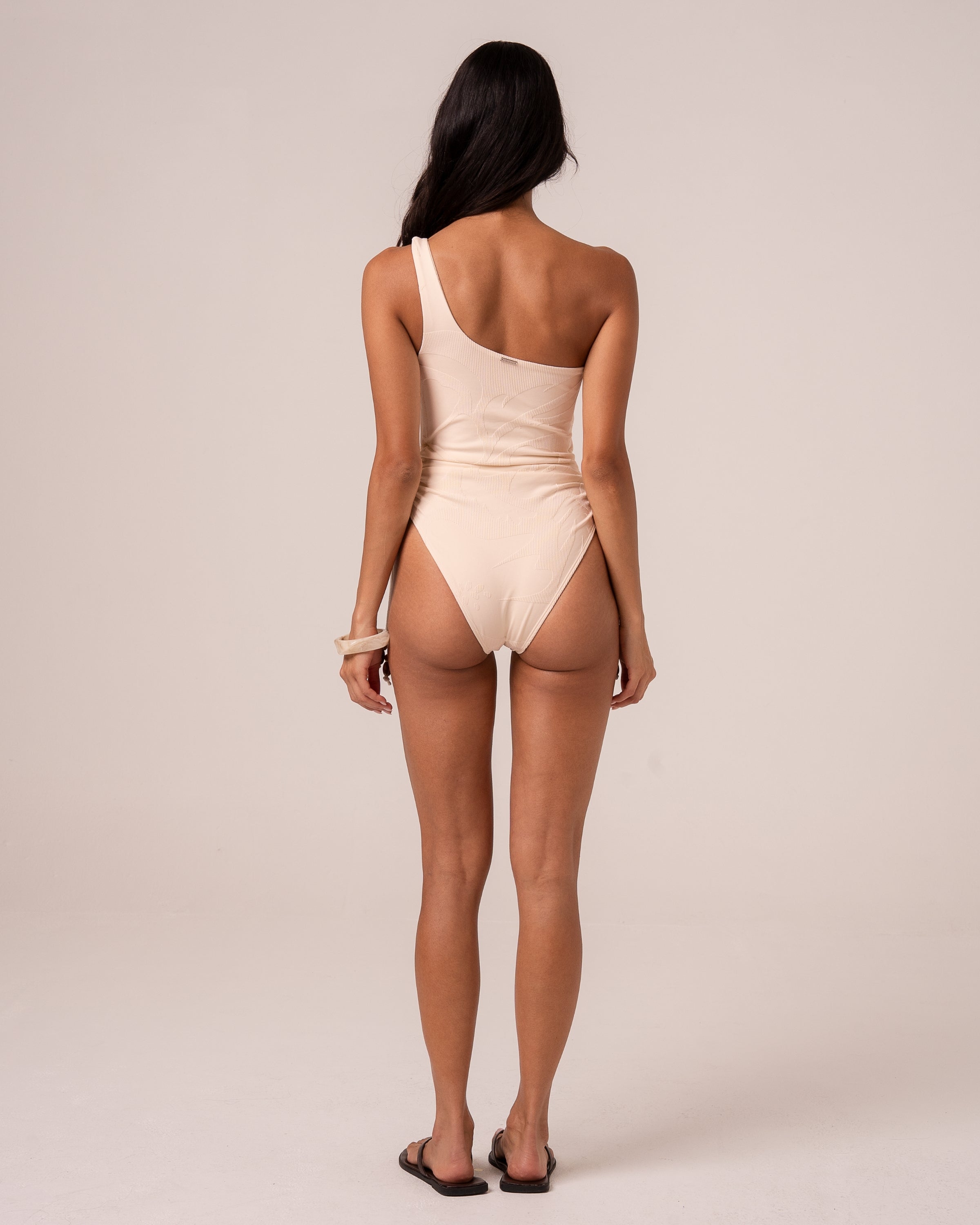 Back view of an ivory asym one shoulder one piece swimsuit showing the single shoulder strap and high-cut leg