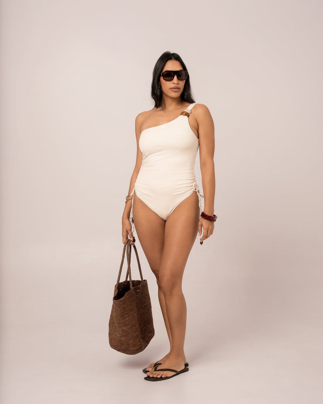 Ivory one shoulder one piece swimsuit styled with a woven raffia tote, showing the adjustable ruched side tie detail — honeymoon resortwear