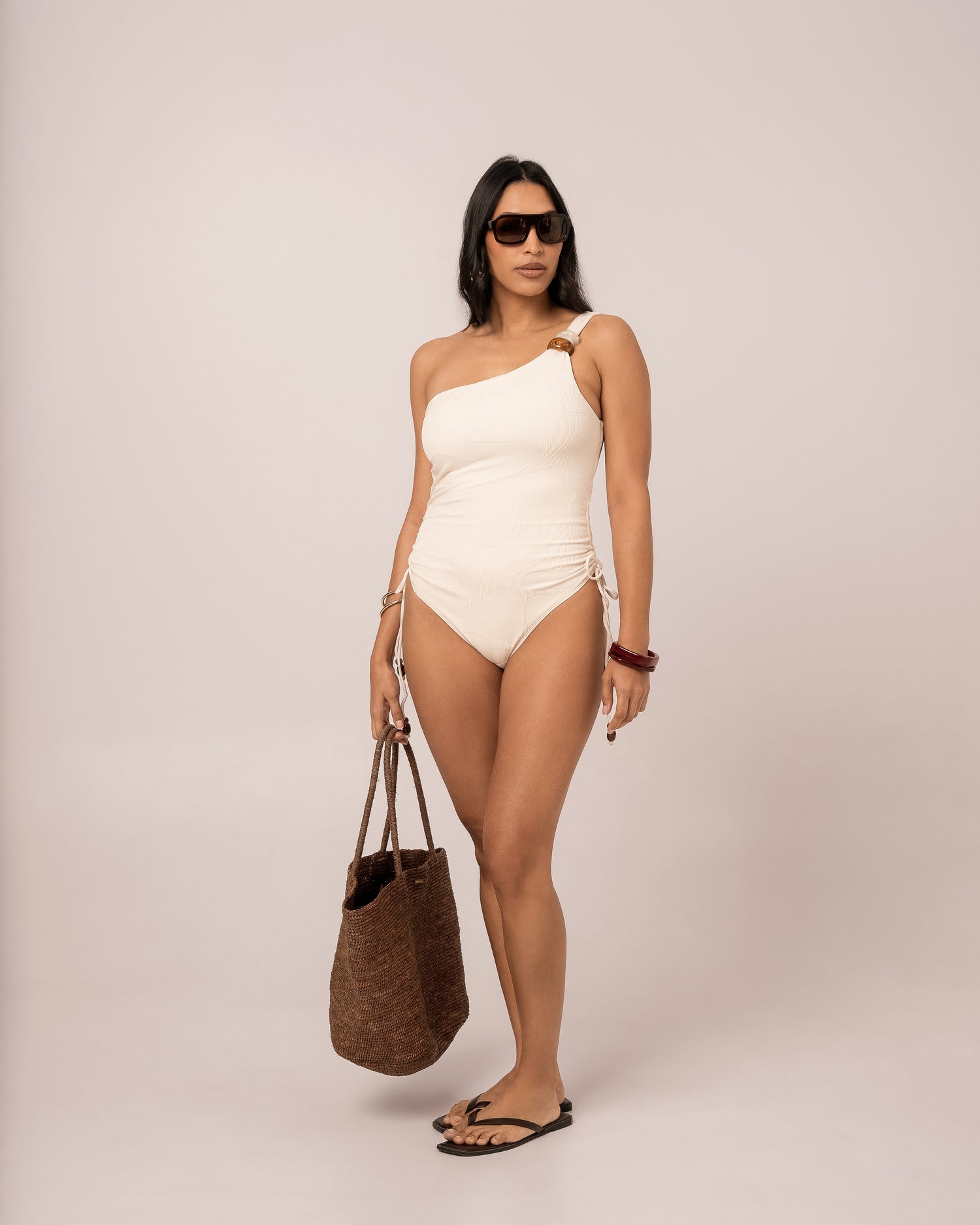 Ivory one shoulder one piece swimsuit styled with a woven raffia tote, showing the adjustable ruched side tie detail — honeymoon resortwear